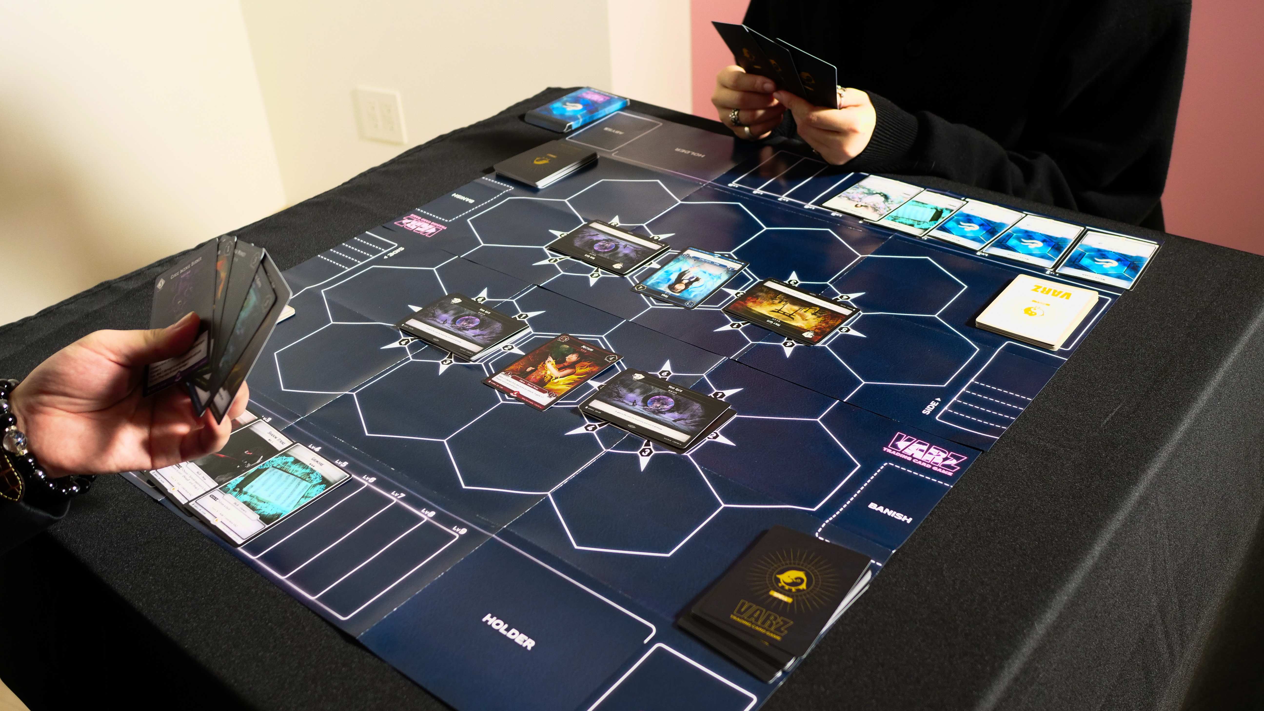 Players in a live VARZ match with starter decks and cards arranged across the hex battlefield playmat
